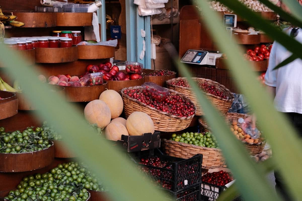 Nahaufnahme eines lebhaften Lebensmittelmarktstandes in Istanbul, gefüllt mit frischem Obst, Gemüse und Gläsern, durch Blätterpflanzen hindurch gesehen.