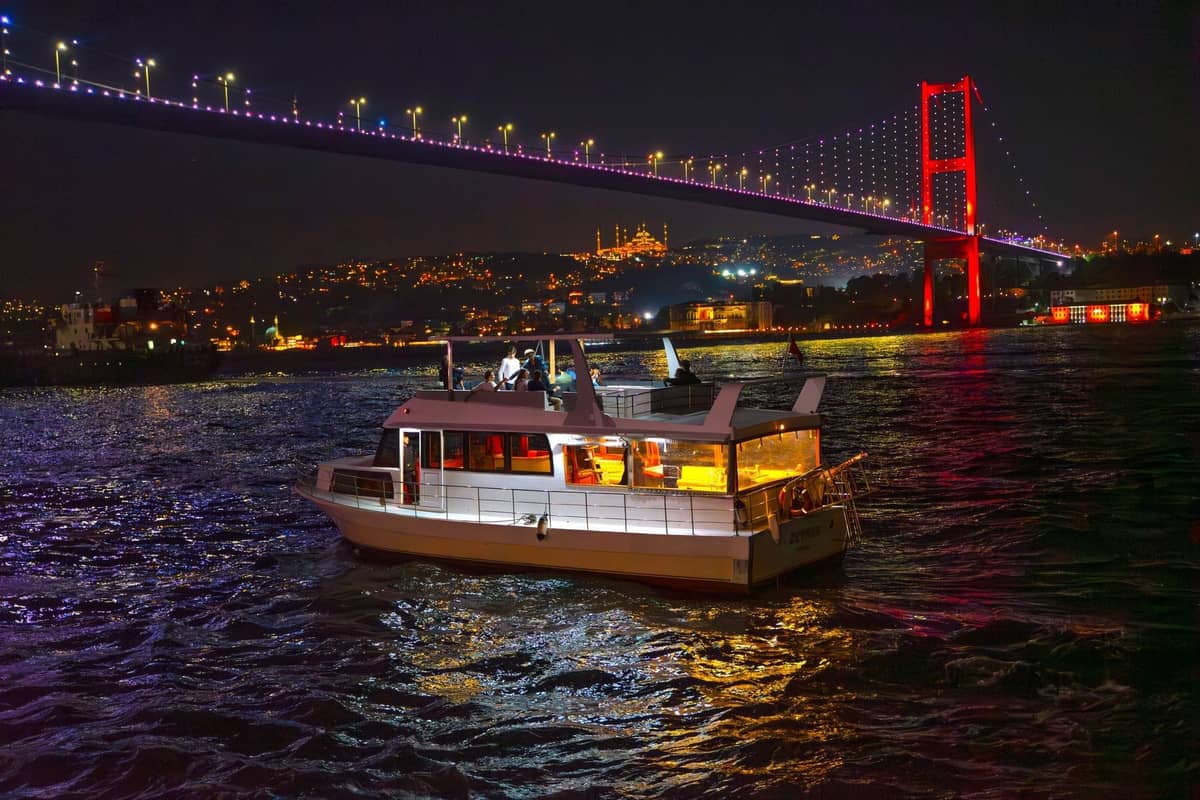 Ein hell erleuchtetes Boot, das nachts auf dem Bosporus fährt, mit der beleuchteten Bosporus-Brücke und der Skyline von Istanbul im Hintergrund.