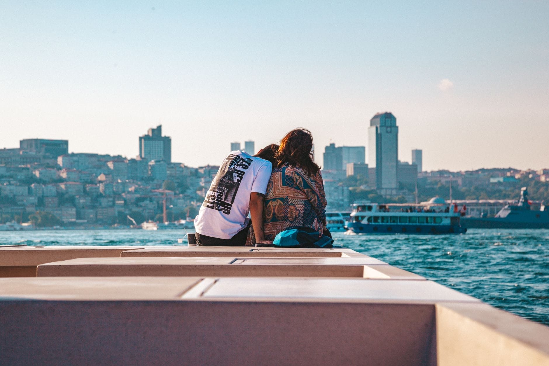 Um casal sentado num banco à beira-mar em Istambul, contemplando o horizonte da cidade e os barcos no Bósforo sob um céu azul intenso.