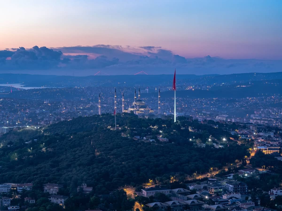 Erhöhte Dämmerungsansicht über einem Hügel Istanbuls mit der Çamlıca-Moschee, der türkischen Flagge und der Stadtsilhouette bei Sonnenuntergang.