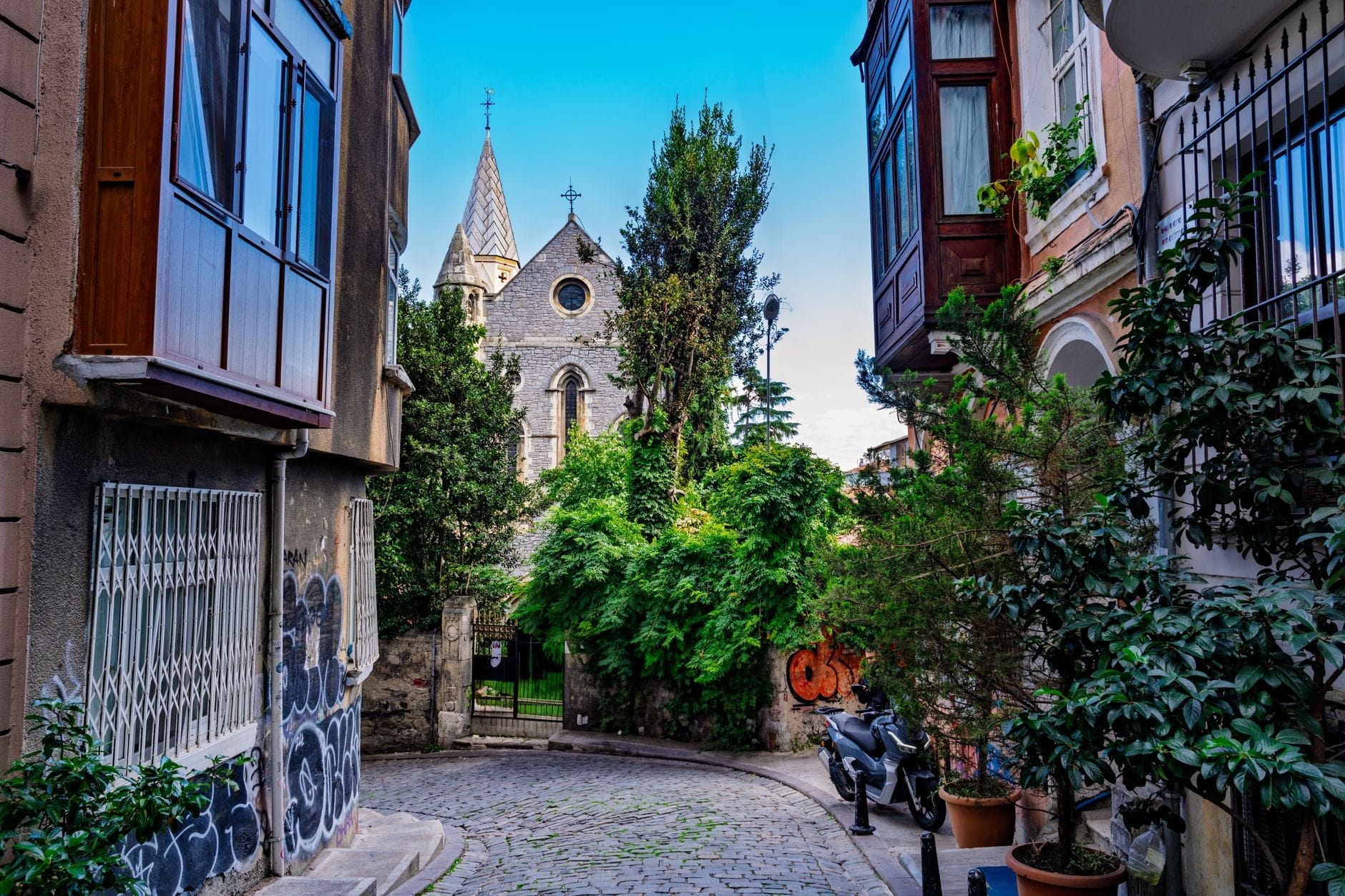 Uma ruazinha de paralelepípedos tranquila em Istambul, ladeada por casas antigas e vegetação, levando a uma igreja de pedra escondida com torres pontiagudas e uma cruz, banhada pela luz vibrante do dia.