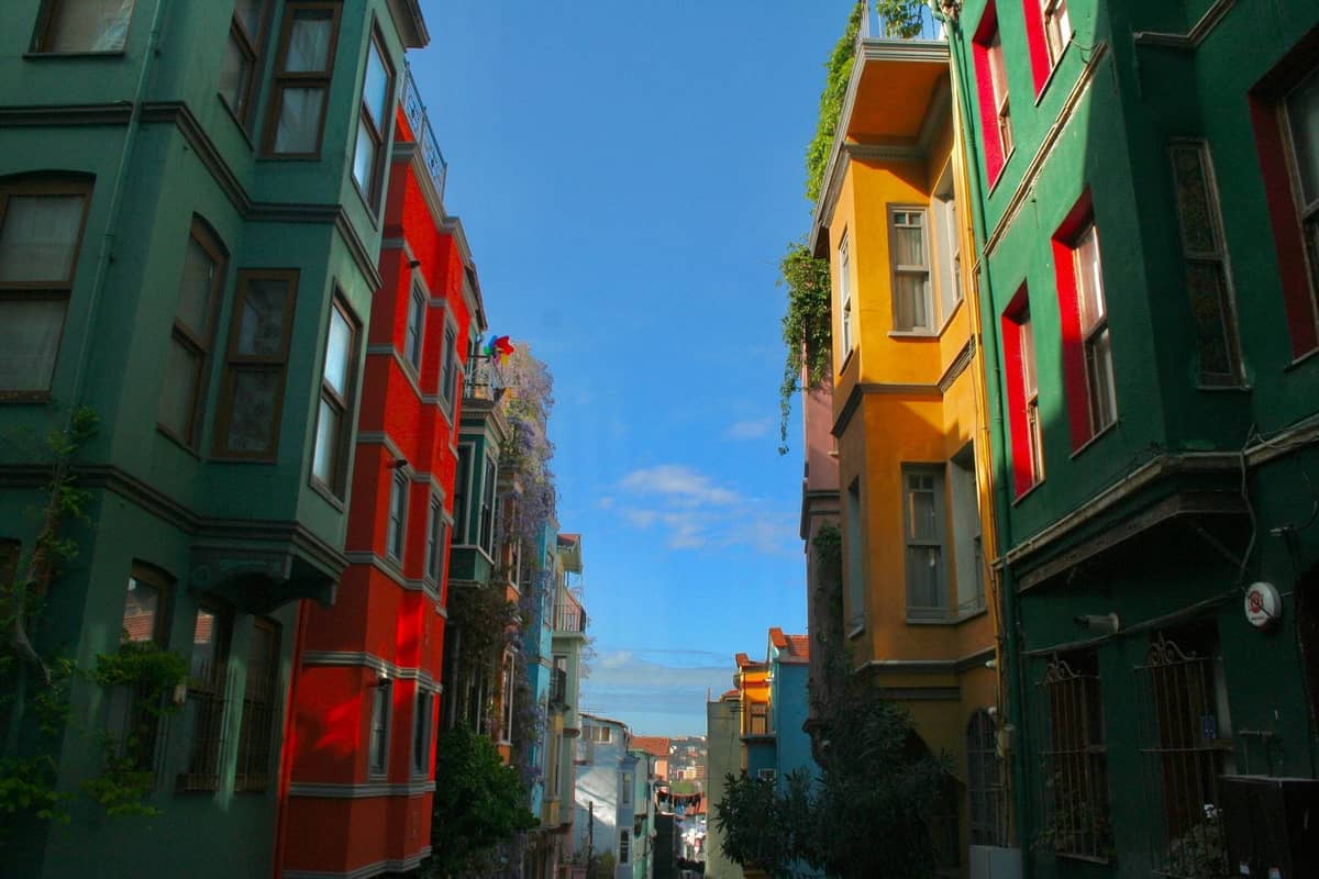 Her iki yanında renkli eski apartman binaları sıralanan, üzerinde mavi gökyüzünün uzandığı ve şehre doğru hafif eğimli bir yokuşa bakan canlı bir İstanbul sokağı.