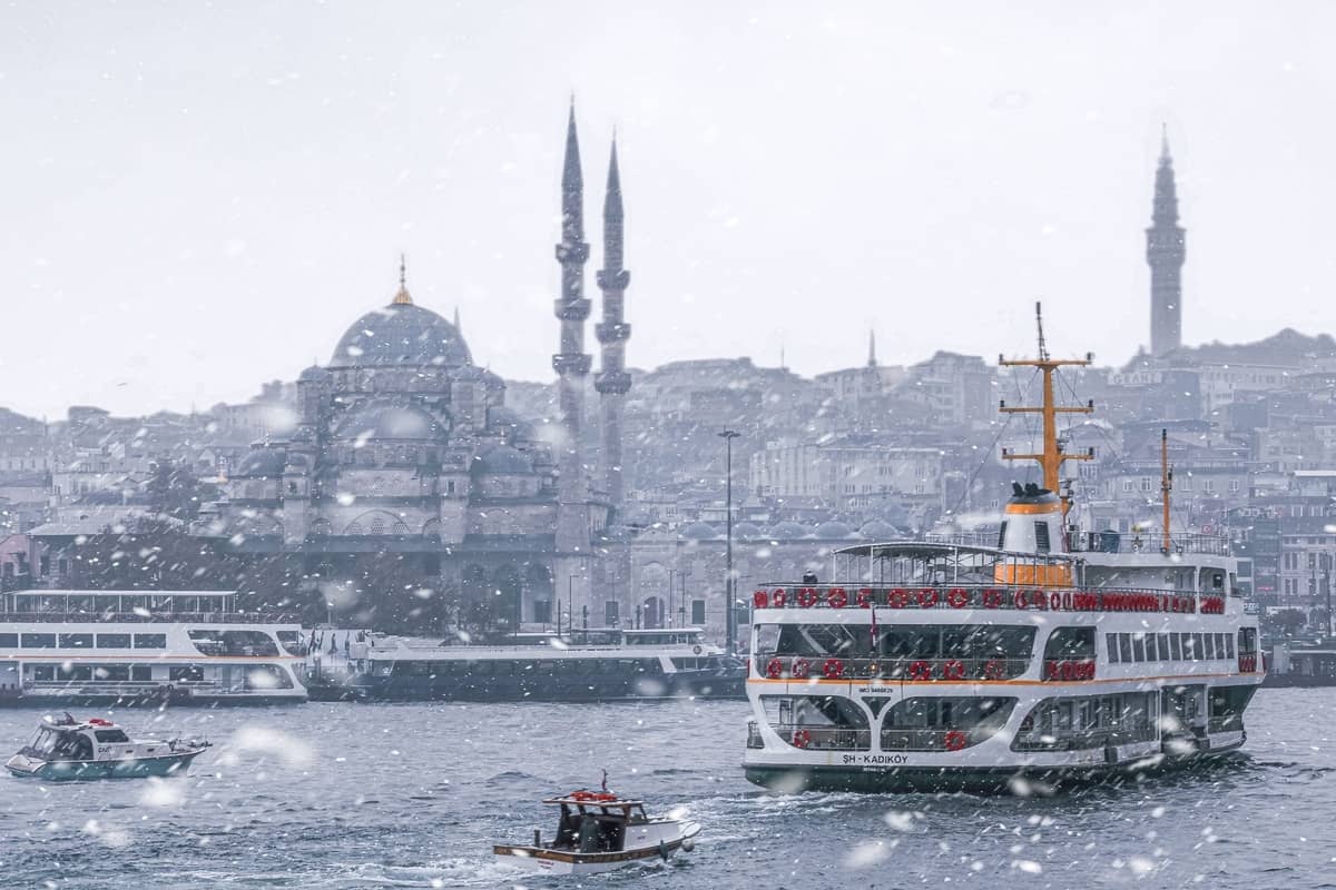 Kış kar yağışında cami ve şehir siluetiyle İstanbul Boğazı'ndaki feribotlar ve tekneler, soğuk havada toplu taşımayı örnekliyor.