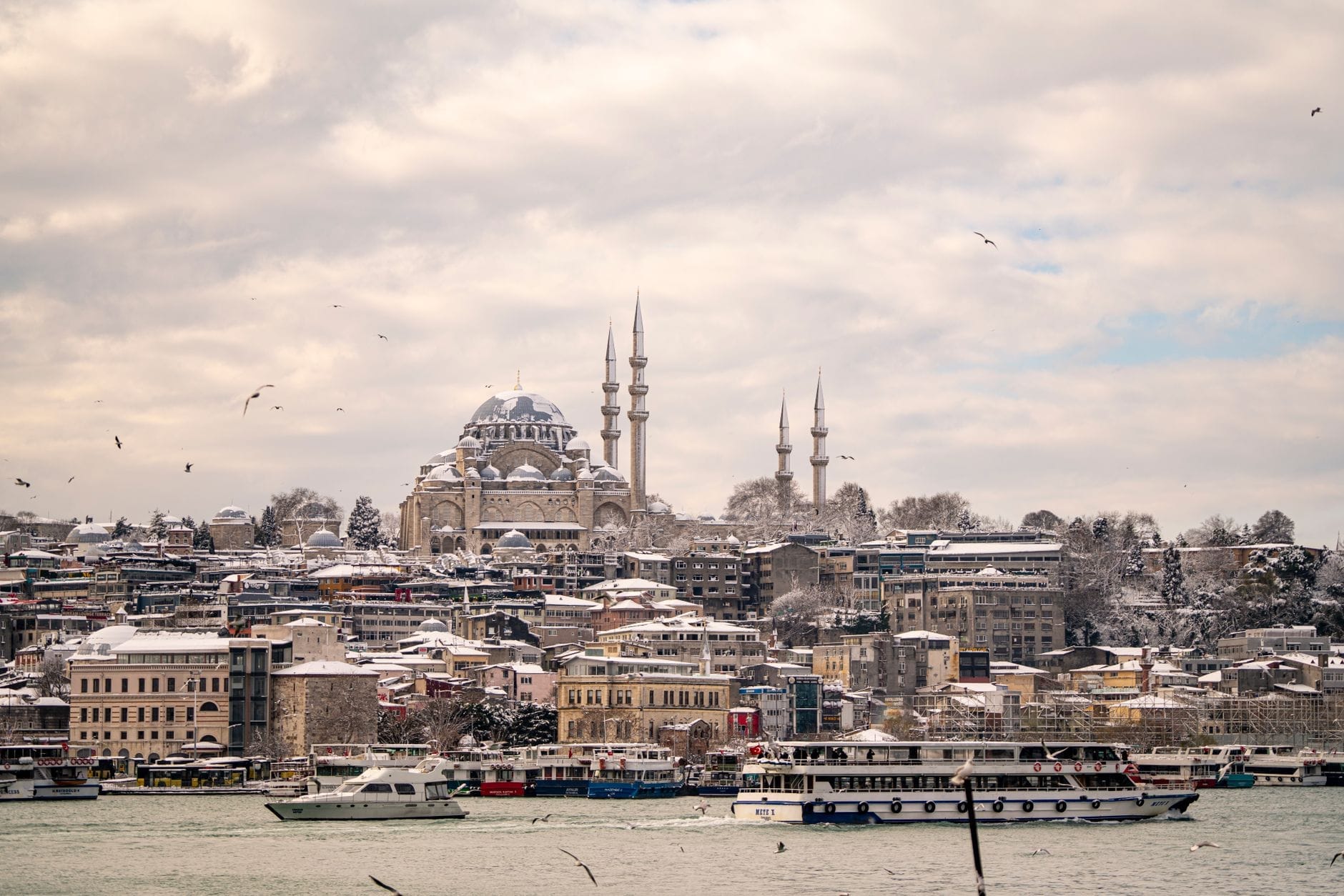 Vista panorâmica de Istambul no inverno com mesquitas históricas, telhados cobertos de neve, balsas no Bósforo e gaivotas sob um céu dramático e suave.