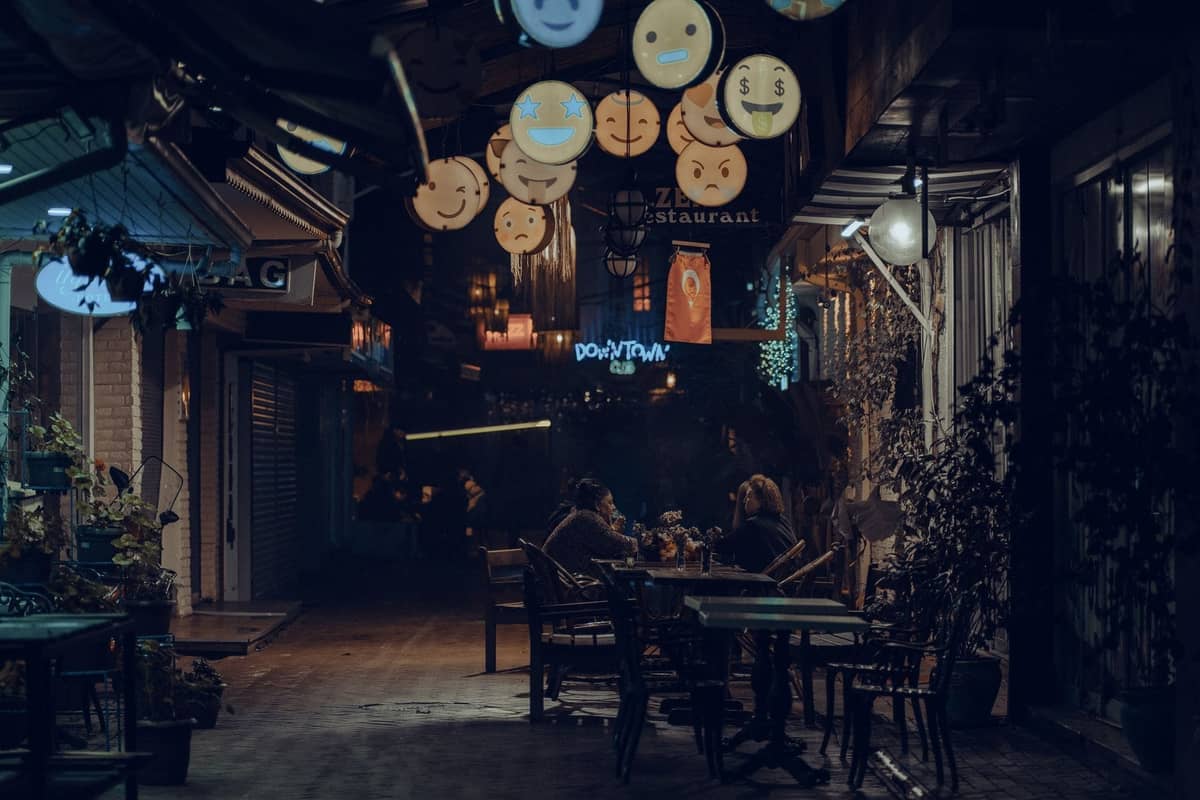 Abendliche Außenszene mit speisenden Menschen an Tischen entlang einer engen, laternenbeleuchteten Gasse mit türkischen Schildern über ihnen.