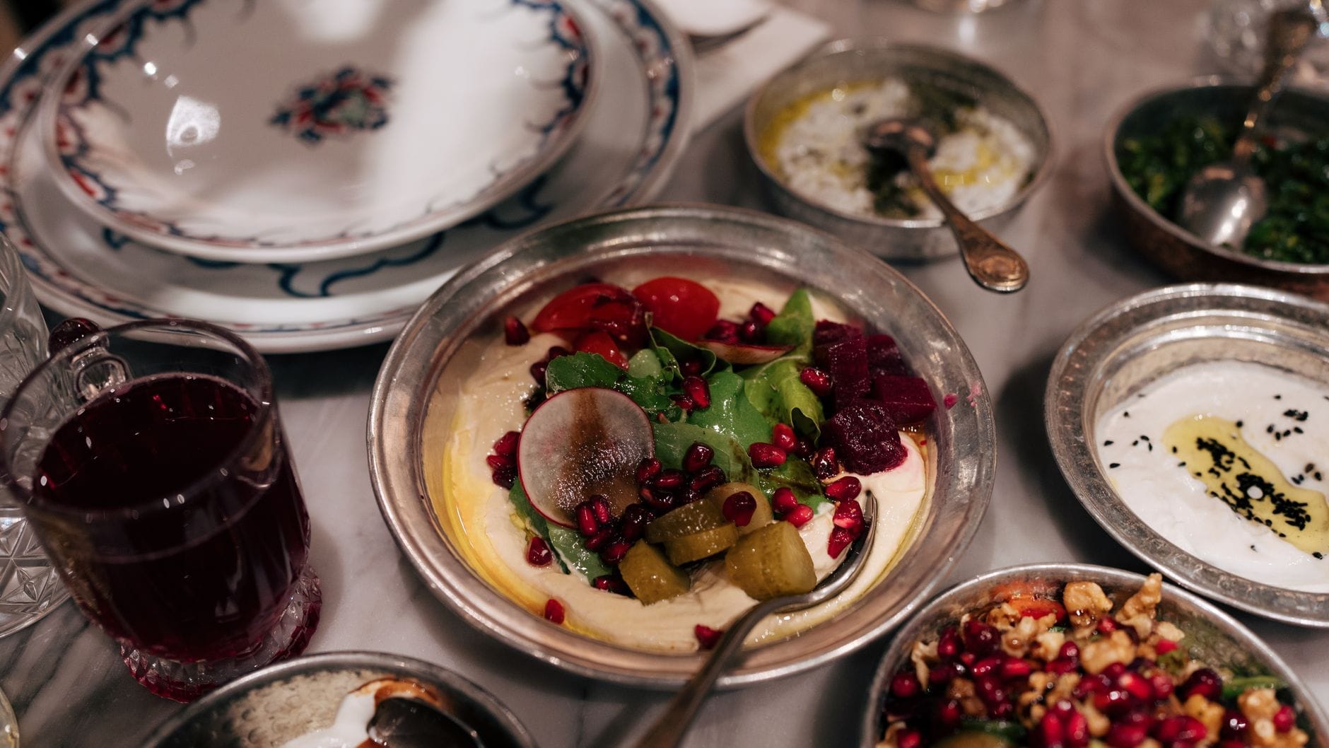 Uma mesa de mármore com tigelas de metal coloridas cheias de pratos de meze variados, com legumes, sementes de romã e picles, evocando o ambiente autêntico de uma meyhane turca.