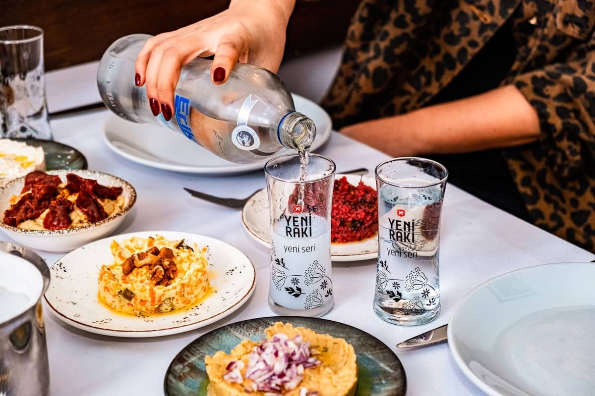 Gedeckter Tisch mit klassischen türkischen Meze-Tellern und zwei Gläsern Rakı, mit einer Person, die Wasser einschenkt, in einer typischen Meyhane-Atmosphäre.
