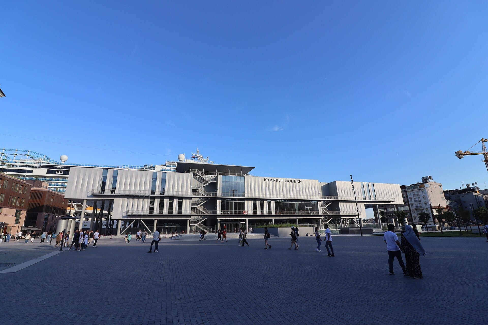 Vista ampia della facciata elegante e moderna del museo Istanbul Modern con persone che camminano sulla spaziosa piazza sotto un cielo azzurro limpido.