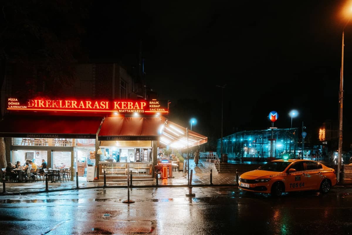 Un ristorante di kebab con un'insegna al neon luminosa e un taxi giallo fuori in una notte piovosa a Istanbul, con l'ingresso della metropolitana visibile.