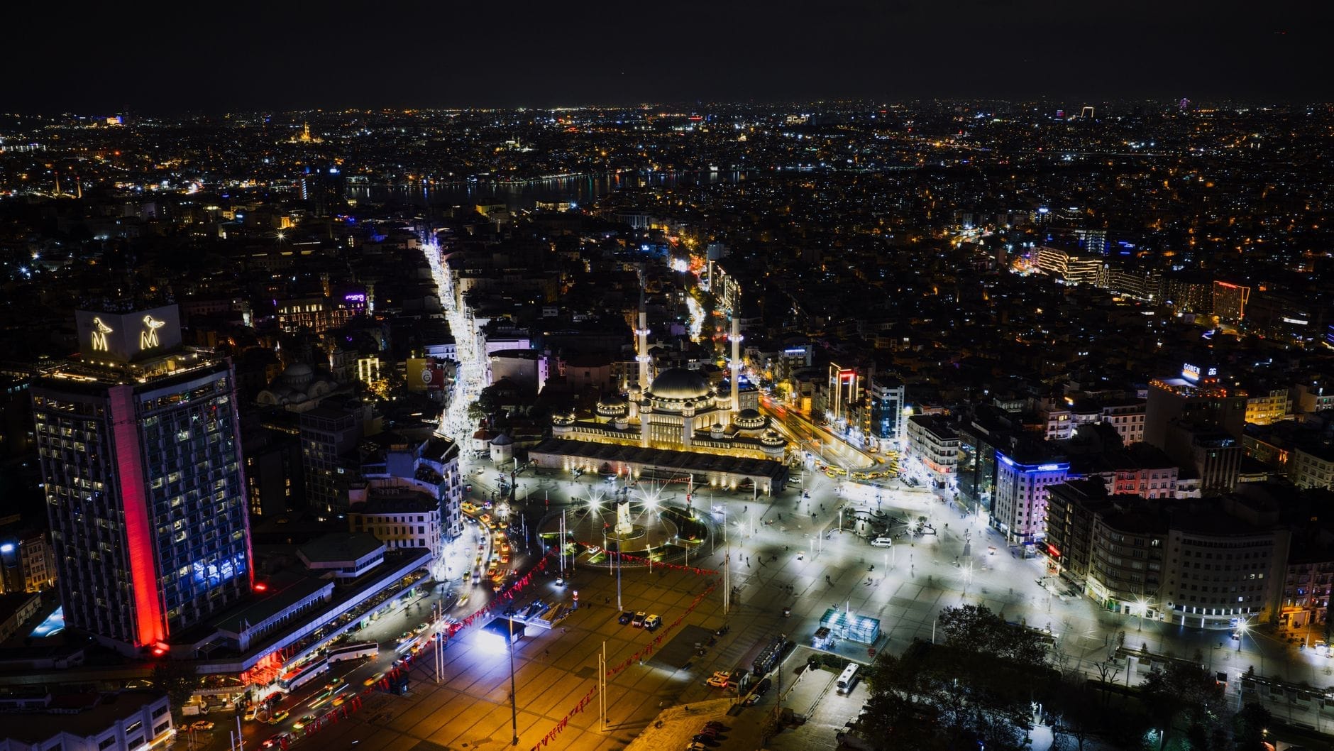 Vista aérea noturna do centro de Istambul com ruas iluminadas, prédios animados e a movimentada Praça Taksim sob uma vasta paisagem urbana que se estende ao horizonte.