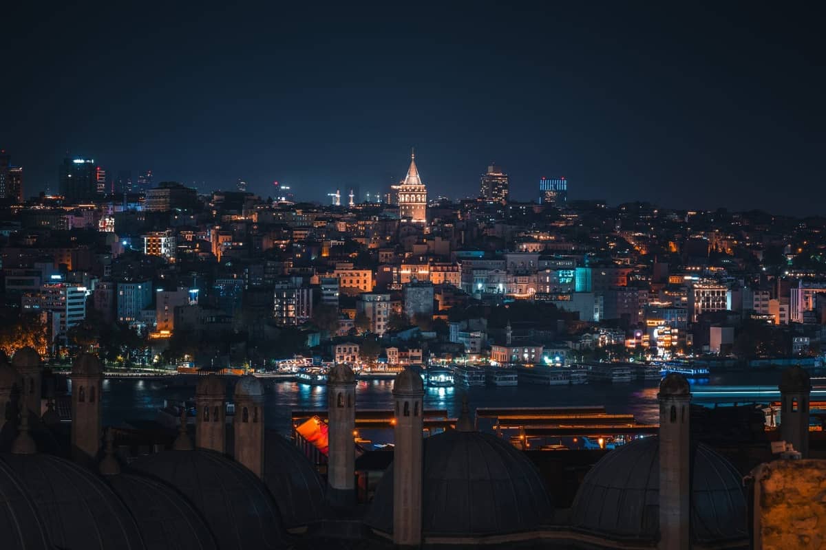 Vista notturna di Istanbul con la Torre di Galata illuminata, le luci della città e cupole sui tetti in primo piano.