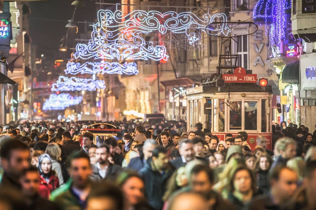 L'affollata Istiklal Avenue di notte con luci festive, un tram nostalgico rosso e folla di persone che godono della vivace vita notturna di Istanbul.