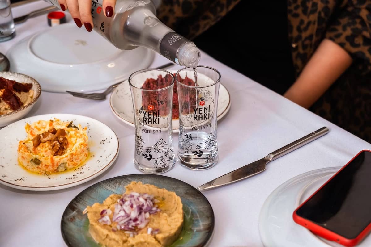 Tavolo con bicchieri di raki, piatti di meze freddi e qualcuno che versa il raki, tipico di un meyhane di İstanbul.