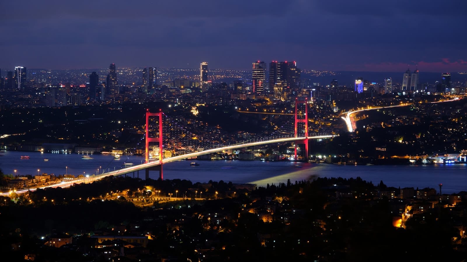 Nighttime panoramic view of Istanbul with the illuminated Bosphorus Bridge spanning across the water and the city skyline in the background.