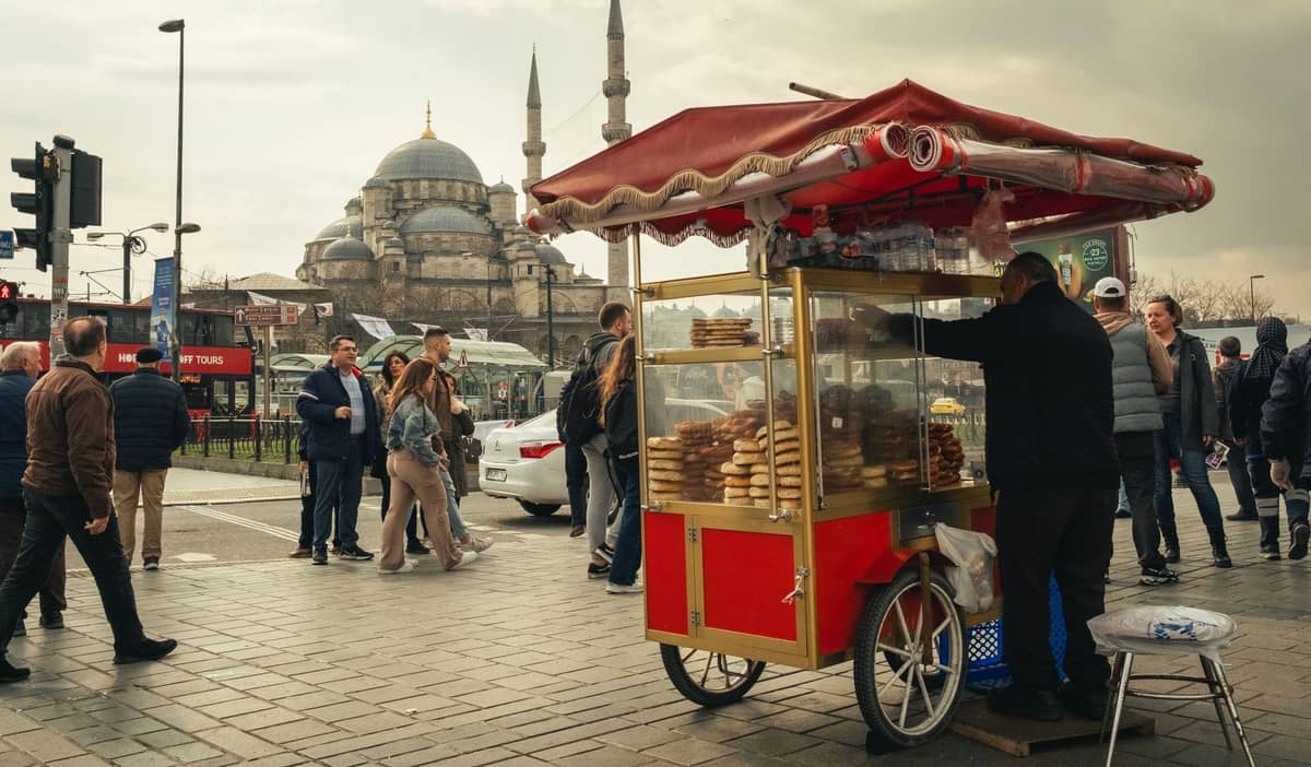 Ein Simit-Straßenverkäufer mit einem roten Wagen, ein belebter Platz in Istanbul und die Kuppeln und Minarette einer Moschee im Hintergrund.