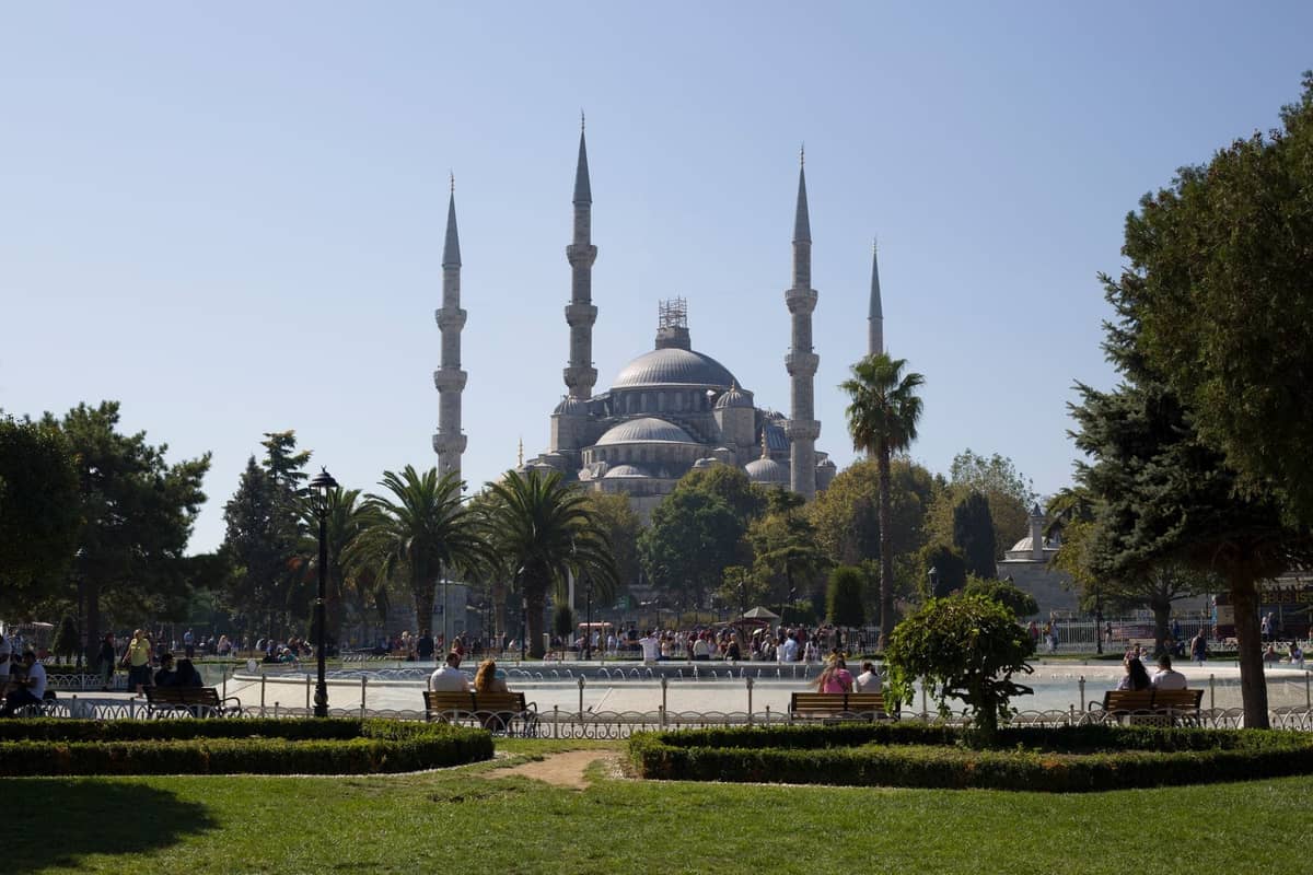 Weitwinkelansicht der Blauen Moschee in Istanbul mit sechs Minaretten und Gärten davor, Menschen auf Parkbänken im Vordergrund, sonniger Tag.
