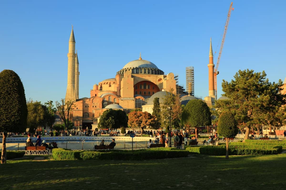 Die Hagia Sophia in Istanbul erhebt sich hinter einem belebten Platz mit Gärten, Bäumen und einem großen Brunnen an einem klaren, sonnigen Tag.