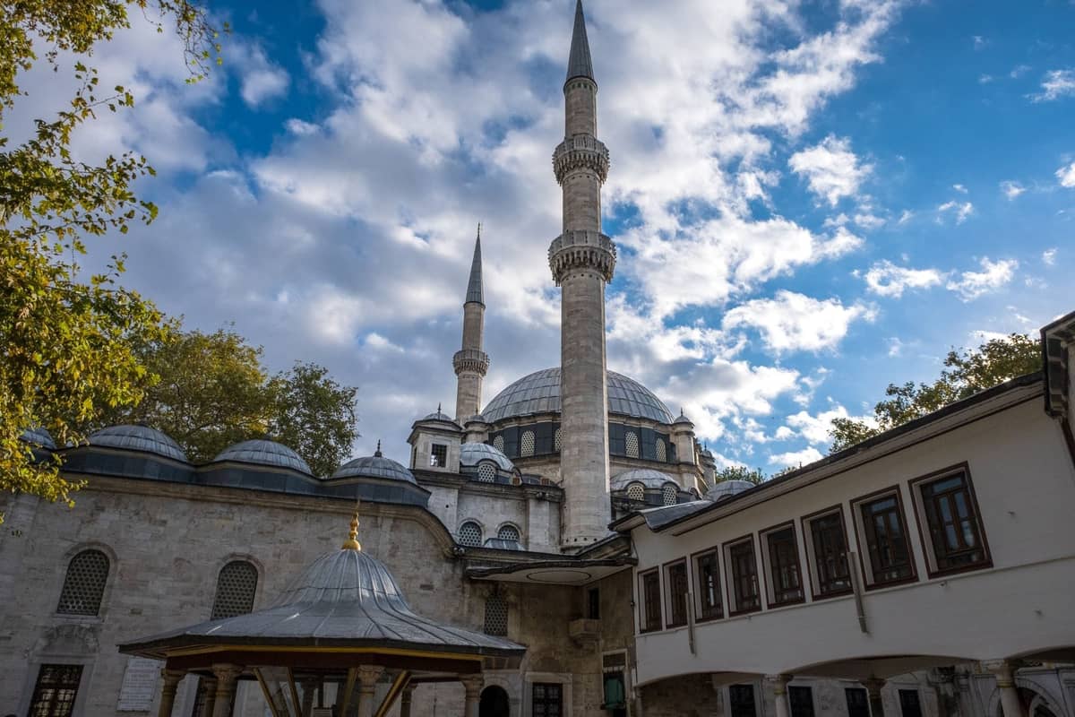 Widok na dziedziniec wielkiego osmańskiego meczetu w Stambule z dwoma wysokimi minaretami na tle częściowo zachmurzonego nieba.