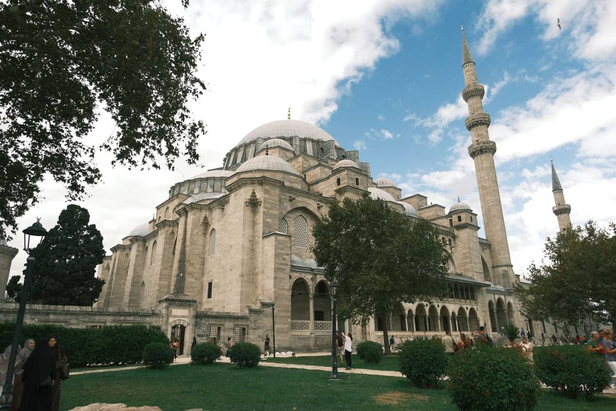 Wielki meczet osmański z kopułami i minaretami, otoczony drzewami i ludźmi, ukazujący klasyczną architekturę Stambułu pod częściowo zachmurzonym niebem.