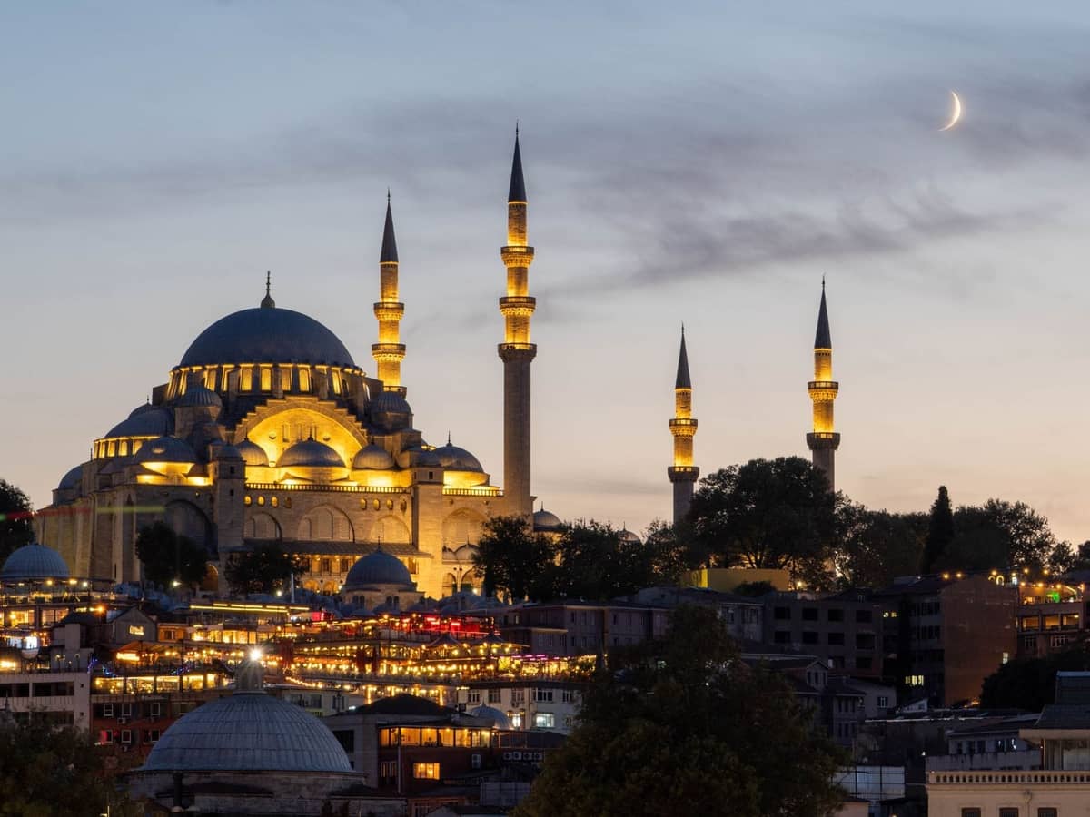 Oświetlony meczet z minaretami w Stambule o zmierzchu, sierp księżyca powyżej, żywe światła miasta poniżej, bezchmurne wieczorne niebo.