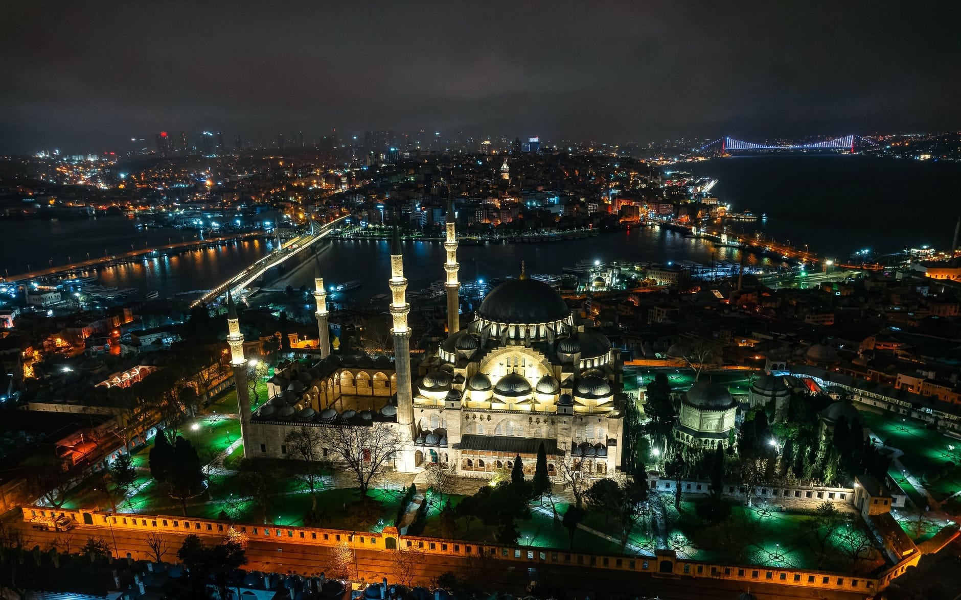 Vista aérea noturna impressionante de uma grande mesquita em Istambul iluminada, com o horizonte da cidade e a Ponte do Bósforo brilhando ao fundo.