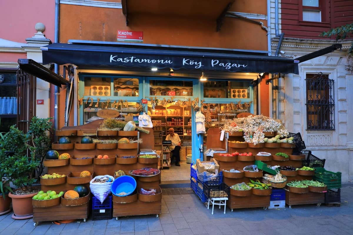 Colorido mercado en Kadıköy con cestas apiladas de productos, un cartel en turco y una persona sentada en el interior. Escena acogedora de compras locales en el lado asiático de Estambul.