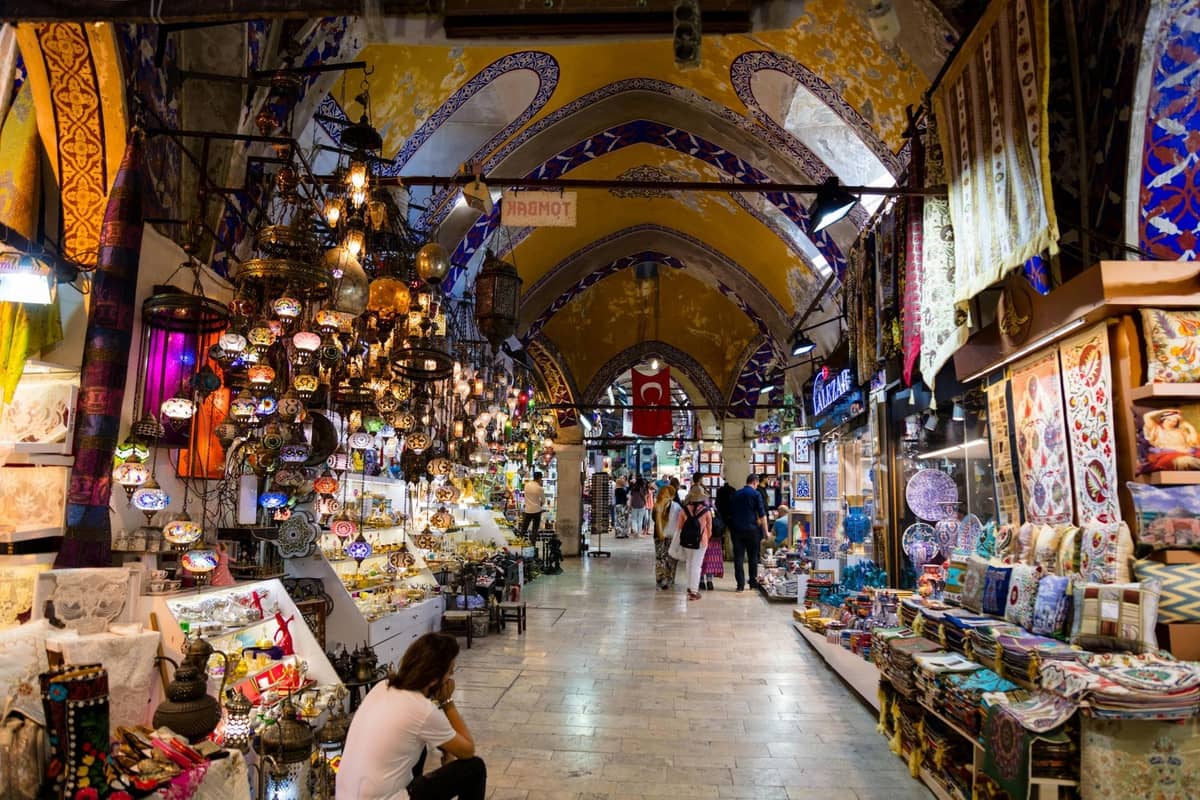 Interior del Gran Bazar de Estambul con techos abovedados, lámparas de colores, textiles y compradores caminando entre puestos concurridos.