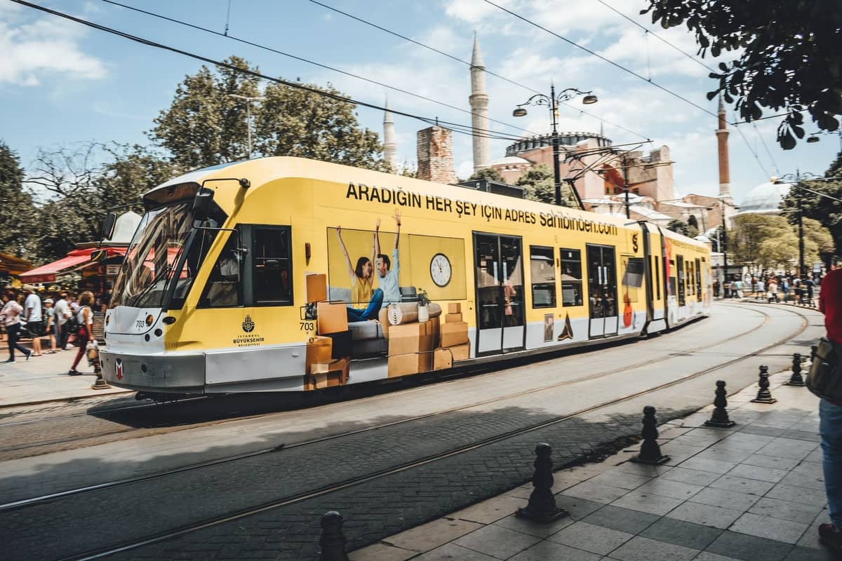 Hellgelbe moderne Straßenbahn, die an der Hagia Sophia vorbeifährt, mit Menschen, die an einem sonnigen Tag in Istanbul in der Nähe spazieren.