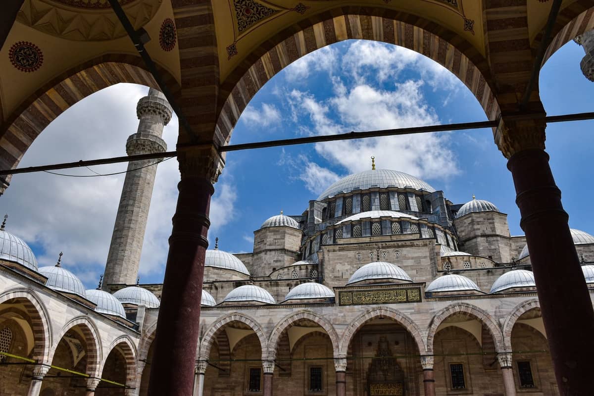 Innenhof und Kuppeln der Süleymaniye-Moschee, von einer gewölbten Kolonnade aus gesehen, mit blauem Himmel und Wolken.