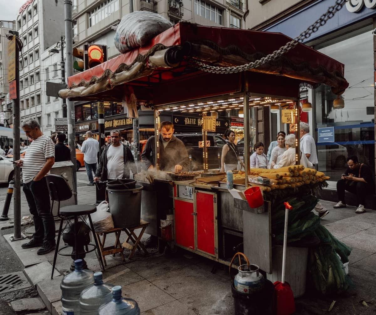 Carrito de comida callejera con maíz y castañas, rodeado de personas en una concurrida calle del barrio de Istanbul.
