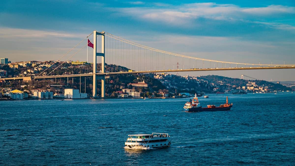 Blick auf die Bosporus-Brücke in Istanbul mit Fähren und Tankern auf dem Wasser, Stadt und Hügel im Hintergrund.