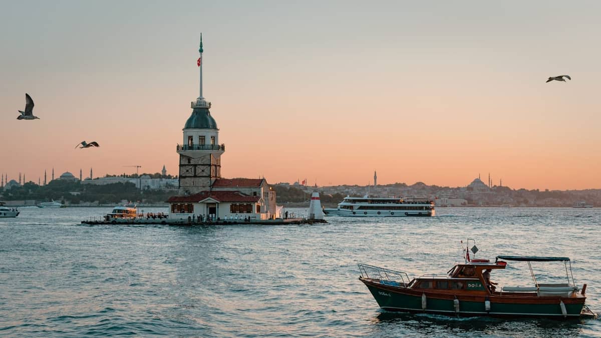 Malerischer Blick auf den Maiden's Tower im Bosporus mit vorbeifahrenden Booten und der Stadtsilhouette bei Sonnenuntergang