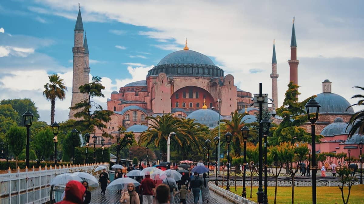 Widok na Hagia Sophia z ludźmi spacerującymi po głównej promenadzie, niektórymi trzymającymi parasole, bujną roślinnością i dramatycznym niebem w Sultanahmet.