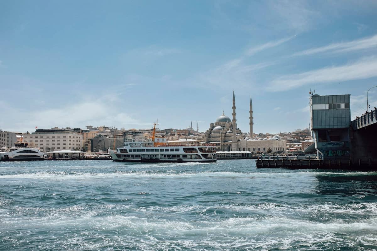 Cena da via fluvial de Istambul com um ferry, o horizonte de mesquitas e edifícios da cidade sob um céu azul limpo, ilustrando um clima ideal para passeios turísticos e atividades náuticas.