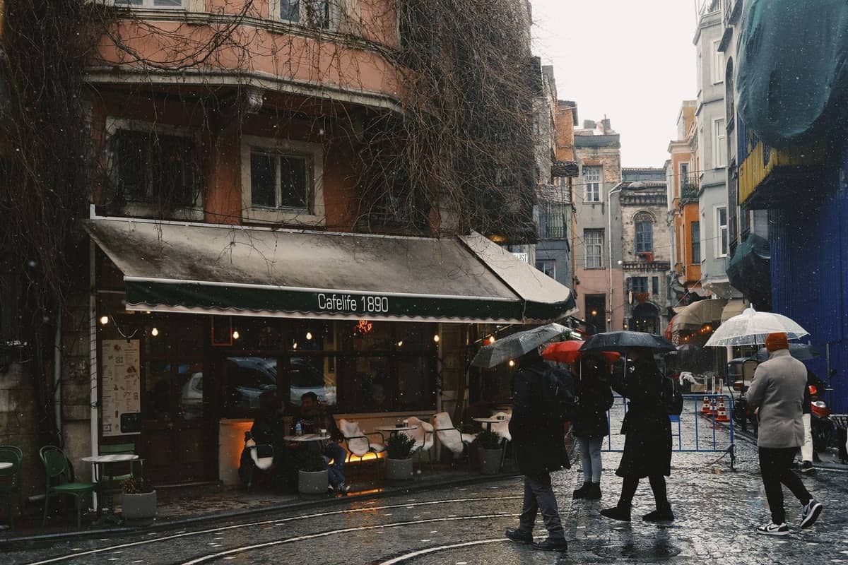 Pessoas caminhando com guarda-chuvas em uma rua molhada em Istambul perto de um café, mostrando o tempo chuvoso e a vida urbana.