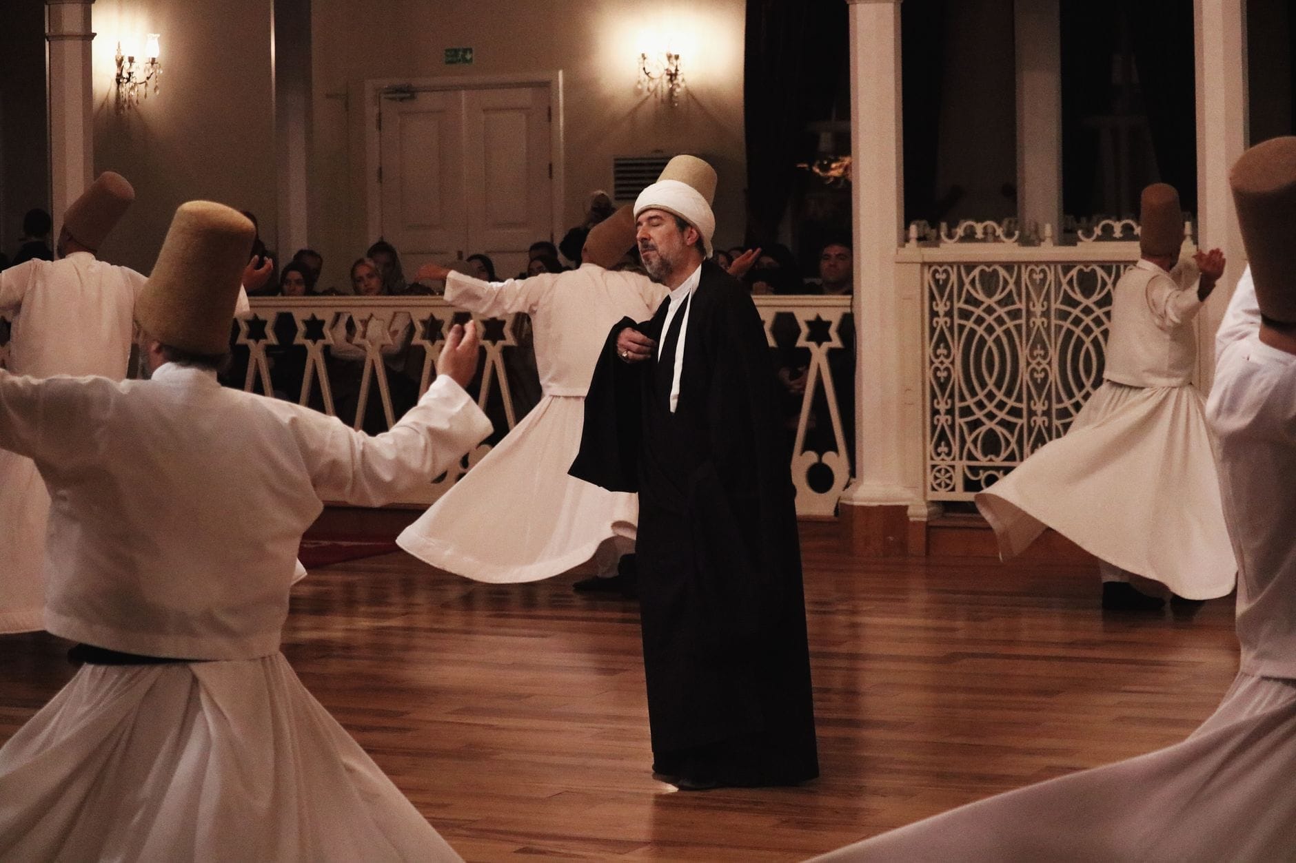 Dervixes giratórios realizando a cerimônia Sema em um salão elegante, vestindo trajes brancos tradicionais e chapéus altos, com espectadores ao fundo.