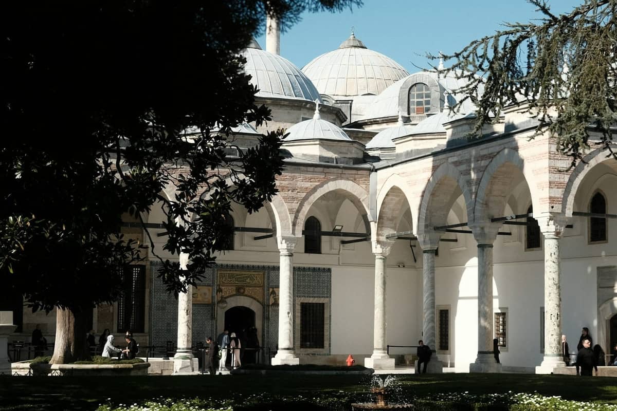 Innenhof des Topkapi-Palastes mit Besuchern, die unter Bäumen spazieren und sitzen, Kuppeldächer und Bögen im Hintergrund an einem sonnigen Tag.