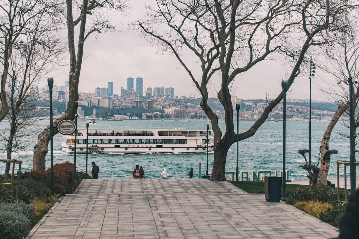 Istanbuler Uferpromenade mit Bäumen, glattем Weg, Bänken und einer Fähre auf dem Bosporus, Stadtsilhouette im Hintergrund.