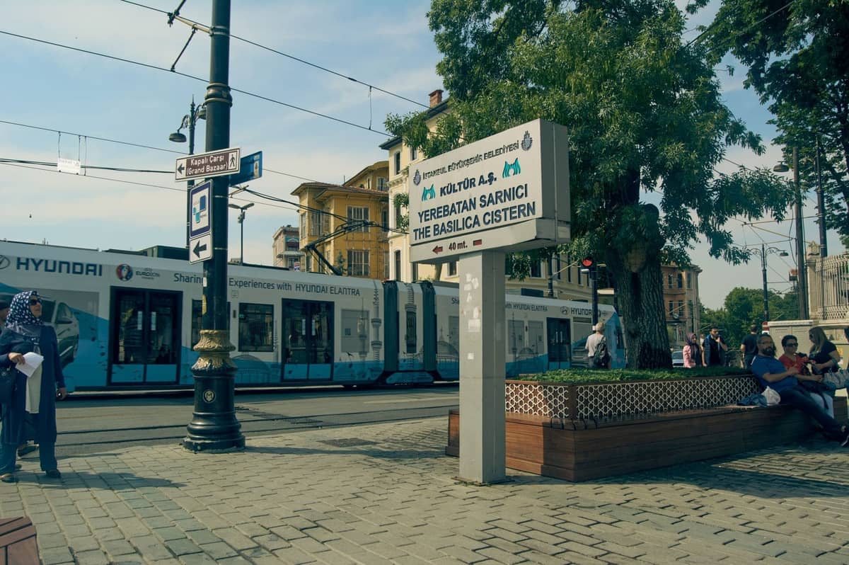 Eine moderne Straßenbahn fährt durch eine zentrale Straße in Istanbul mit gut sichtbaren Hinweisschildern zu Touristenattraktionen und wartenden Menschen in der Nähe.
