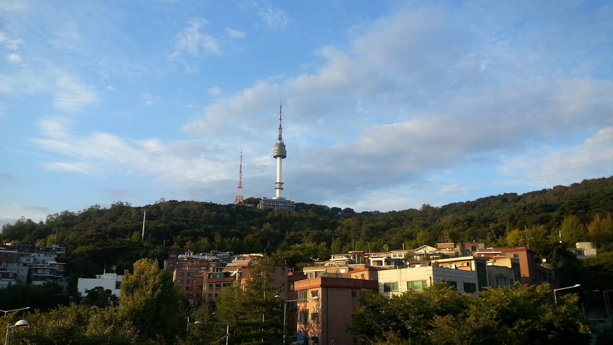 Vista da Torre Namsan no alto do Monte Namsan, ao fundo dos prédios baixos e da vegetação dos bairros de Itaewon e Yongsan, sob um céu azul.