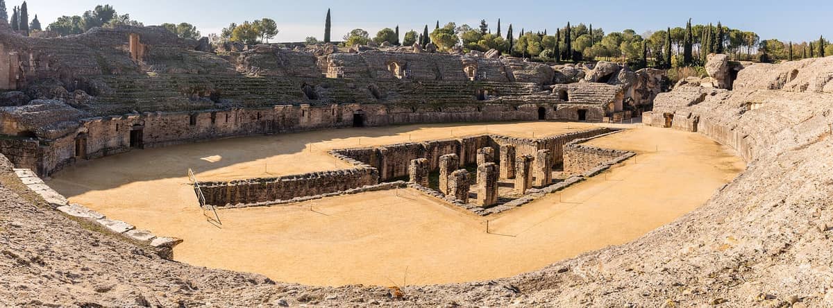 วิวพาโนรามาของอัฒจันทร์โรมันโบราณที่ Itálica แสดงให้เห็นที่นั่งหิน ซากปรักหักพังตรงกลาง และต้นไม้สีเขียวโดยรอบใต้ท้องฟ้าแจ่มใส