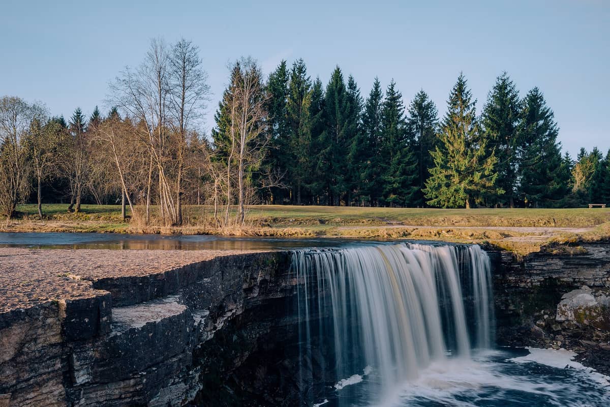 Der Jägala-Wasserfall in Estland: Ein breiter, natürlicher Kalksteinvorsprung, über den Wasser in ein Becken stürzt, umgeben von Bäumen und Grünflächen unter klarem Himmel.