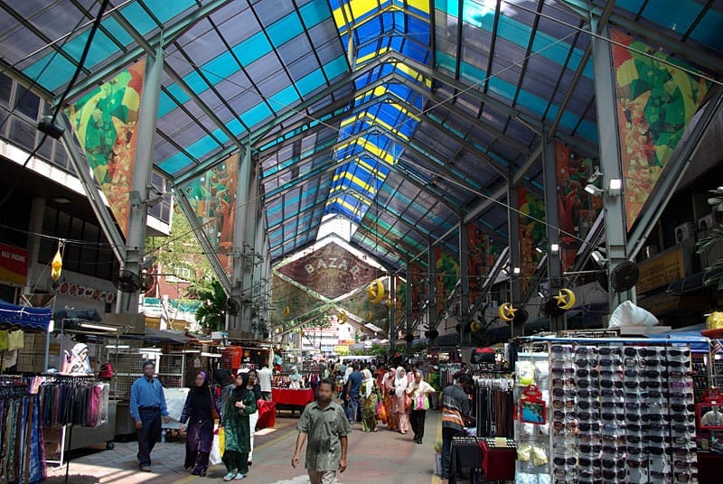 Covered market scene on Jalan Masjid India with colorful glass roof, busy stalls selling textiles and sunglasses, and people walking through the corridor.