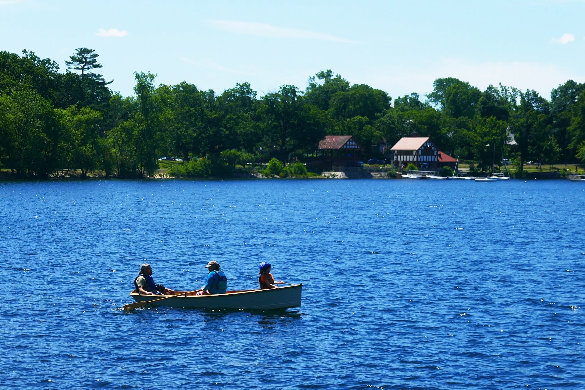 Tre persone remano su una piccola barca sul Jamaica Pond con alberi rigogliosi e gli edifici del rimessaggio sullo sfondo in una giornata serena.