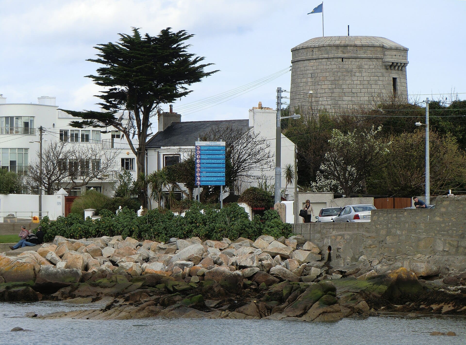 La James Joyce Tower & Museum, una torre Martello rotonda in pietra, si erge lungo rive rocciose, con case moderne, verde e un cartello informativo blu nelle vicinanze.
