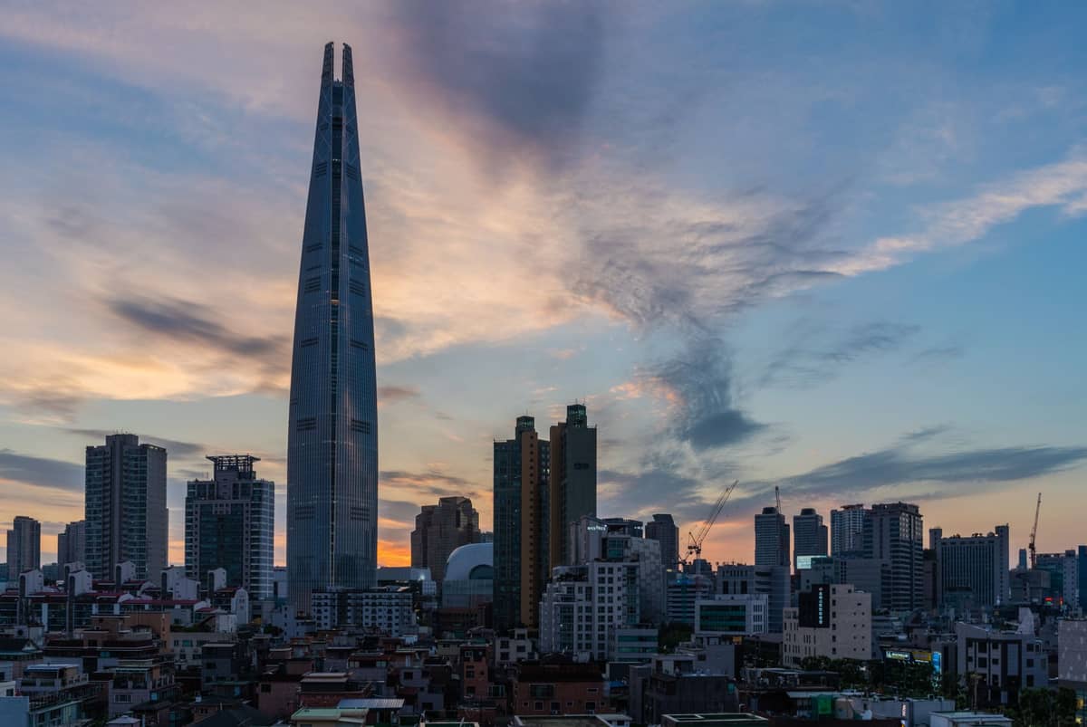 A Lotte World Tower se destacando imponente sobre o skyline de Jamsil e Songpa ao pôr do sol, com arranha-céus modernos ao redor e um céu dramático.