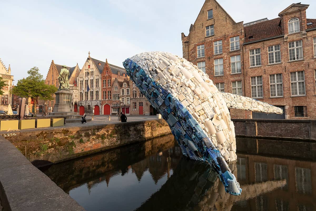 Jan Van Eyckplein in Bruges showing the Jan Van Eyck statue, medieval guild houses, and canal, with a striking sculpture of a whale made from plastic waste in the water.