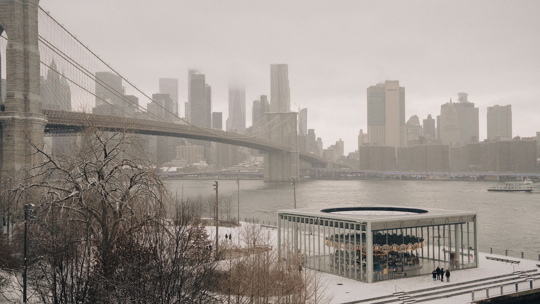 La Giostra di Jane nel suo padiglione di vetro in una giornata di neve, incorniciata dal Brooklyn Bridge e dallo skyline di Manhattan lungo l'East River a Brooklyn Bridge Park.