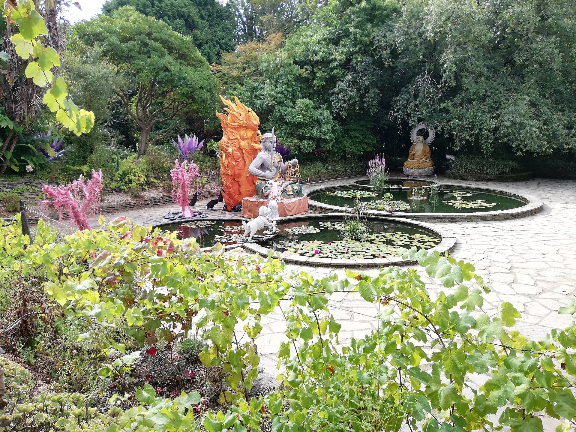 Stagni circolari di ninfee con statue colorate, tra cui un Buddha, circondati da una lussureggiante vegetazione al Jardim Botânico do Porto in una giornata luminosa.