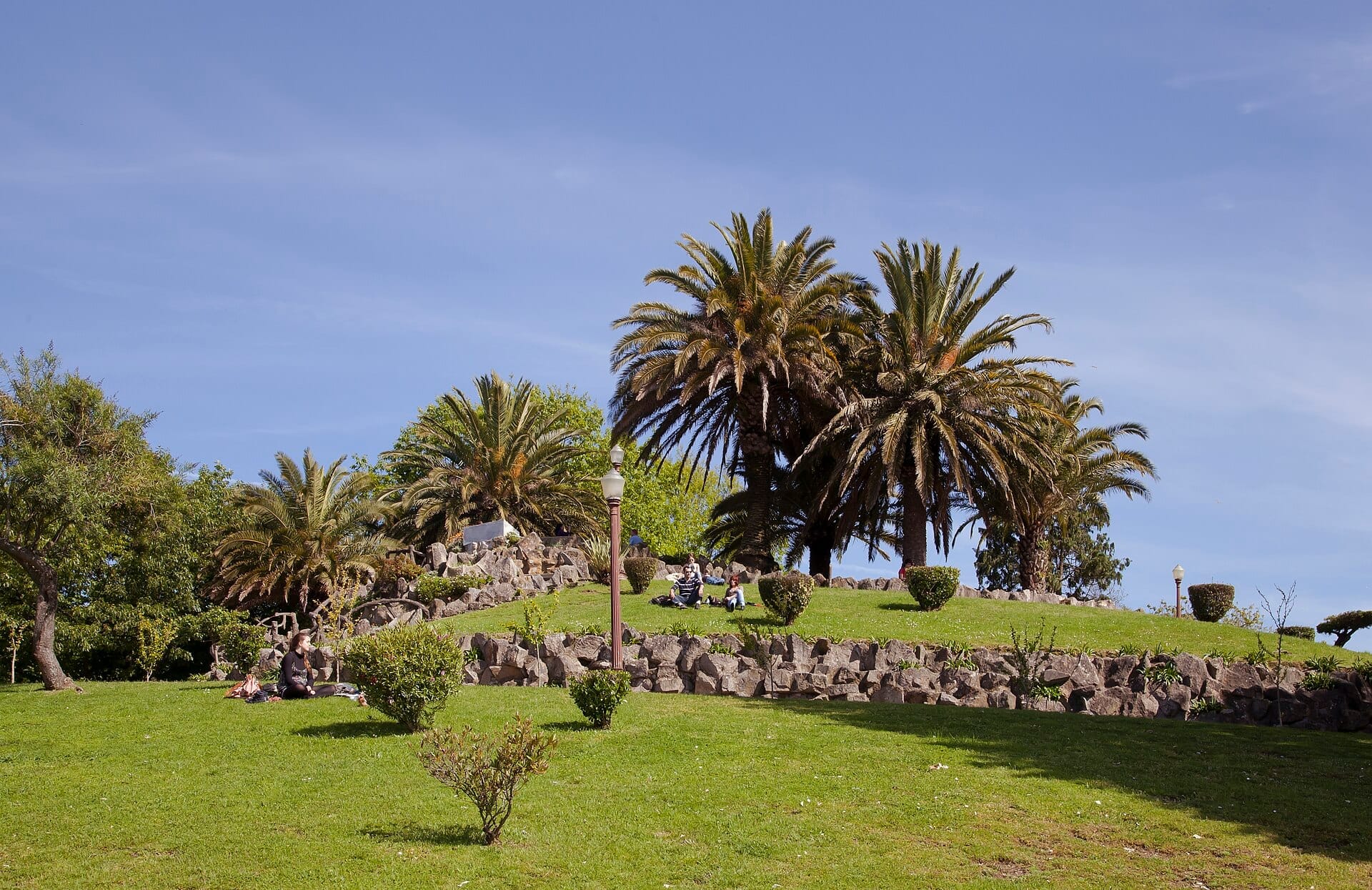 Persone si rilassano sui prati verdi del Jardim do Morro, circondati da palme e terrazze in pietra sotto un cielo azzurro limpido.